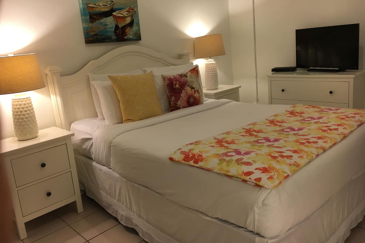A neatly arranged bedroom with a bed, two bedside tables, a lamp, a television, and a painting on the wall.