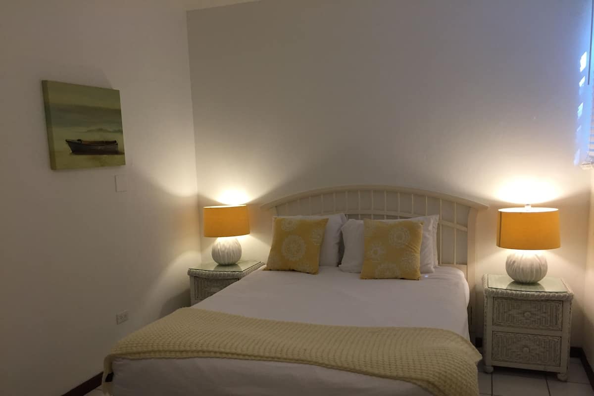 A neatly made bed with a white and yellow bedspread, flanked by two bedside tables with lamps, and a painting on the wall.
