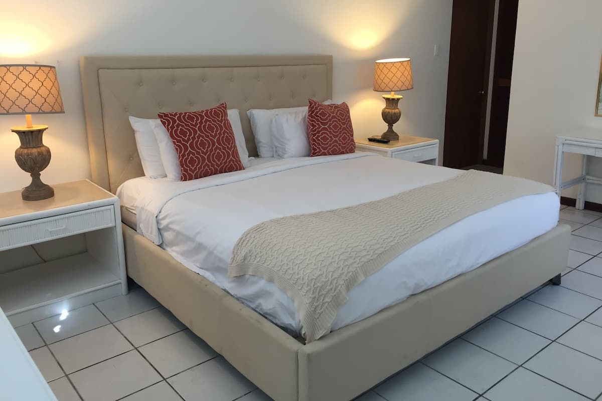 A neatly made bed with a beige headboard, white bedding, and two red patterned pillows. There are two bedside tables with lamps, one on each side of the bed.