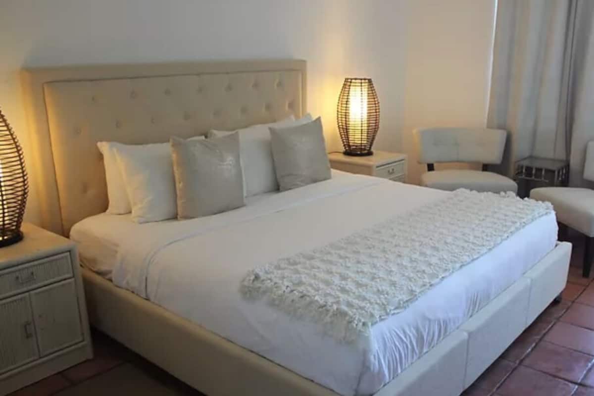 A large bed with a tufted headboard, two bedside tables with lamps, a chair, and a small table.