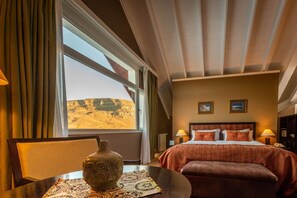 Habitación de hotel con un ventanal grande, cama con ropa de cama roja, mesita de noche con lámpara y un jarrón decorativo.