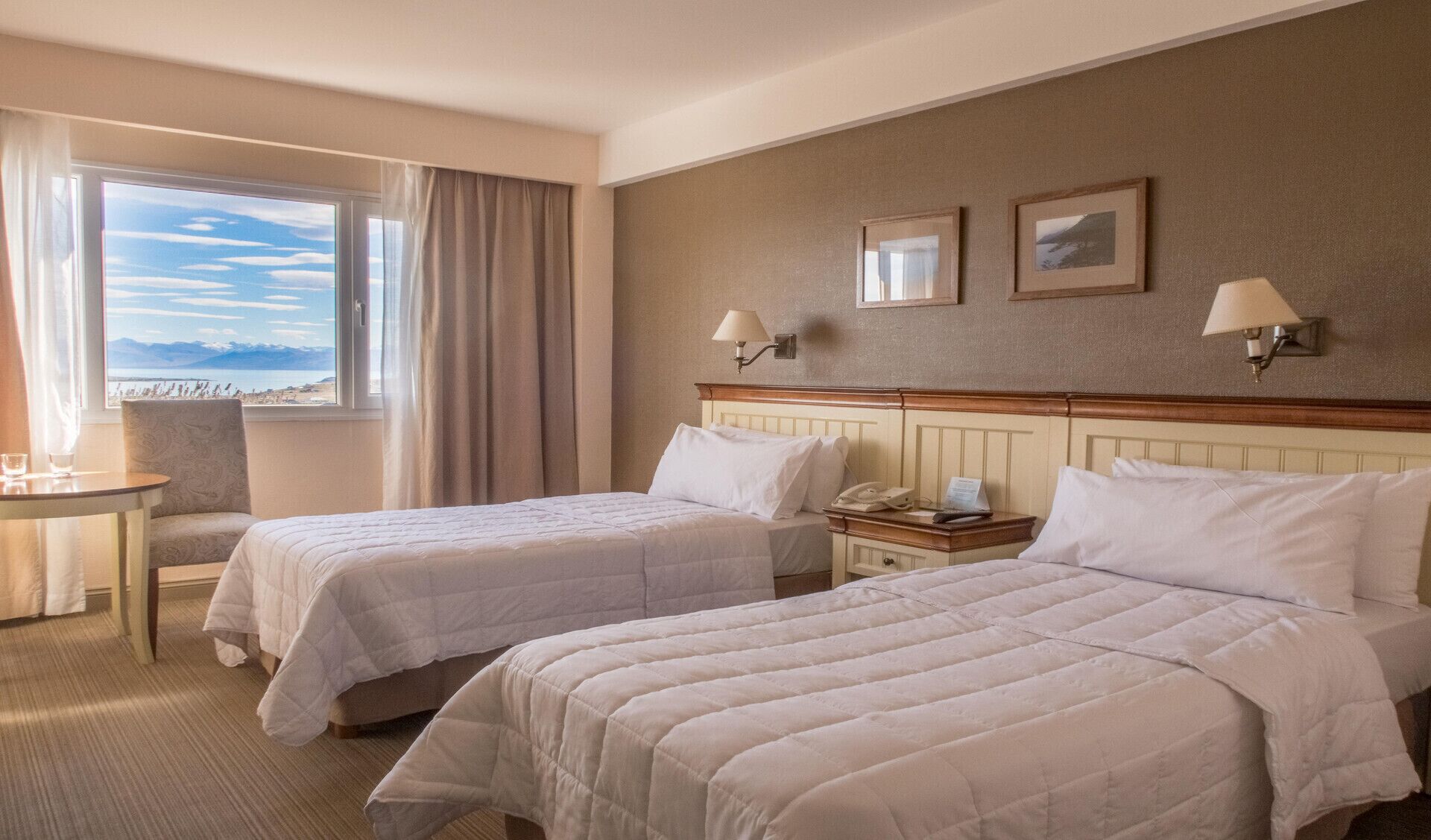 A hotel room with two beds, a desk, a chair, a window with a view, and two wall-mounted lamps.
