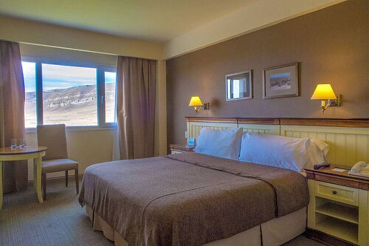 Una camera d'albergo con un letto grande, due lampade ai comodini, un tavolino con una sedia e vista sull'esterno.