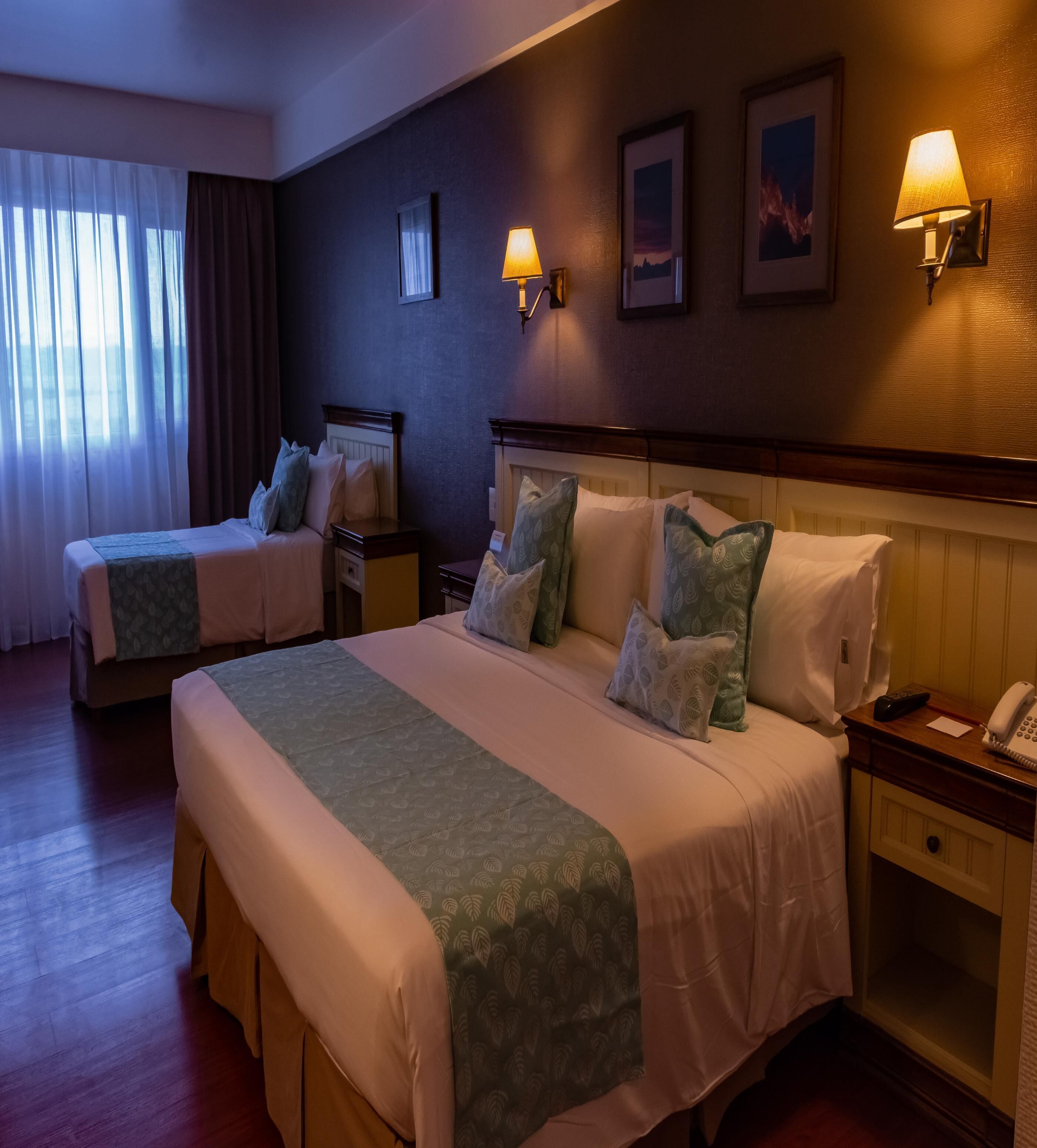 A hotel room with two beds, bedside lamps, a nightstand with a phone, and framed pictures on the wall.