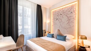 Habitación de hotel con cama, escritorio y silla. Cuenta con un ventanal con cortinas, una lámpara de pared y una obra de arte colgada.