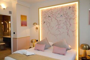 A hotel room with a bed, pillows, a nightstand, a lamp, a wall with a floral mural, and a bathroom visible in the background.