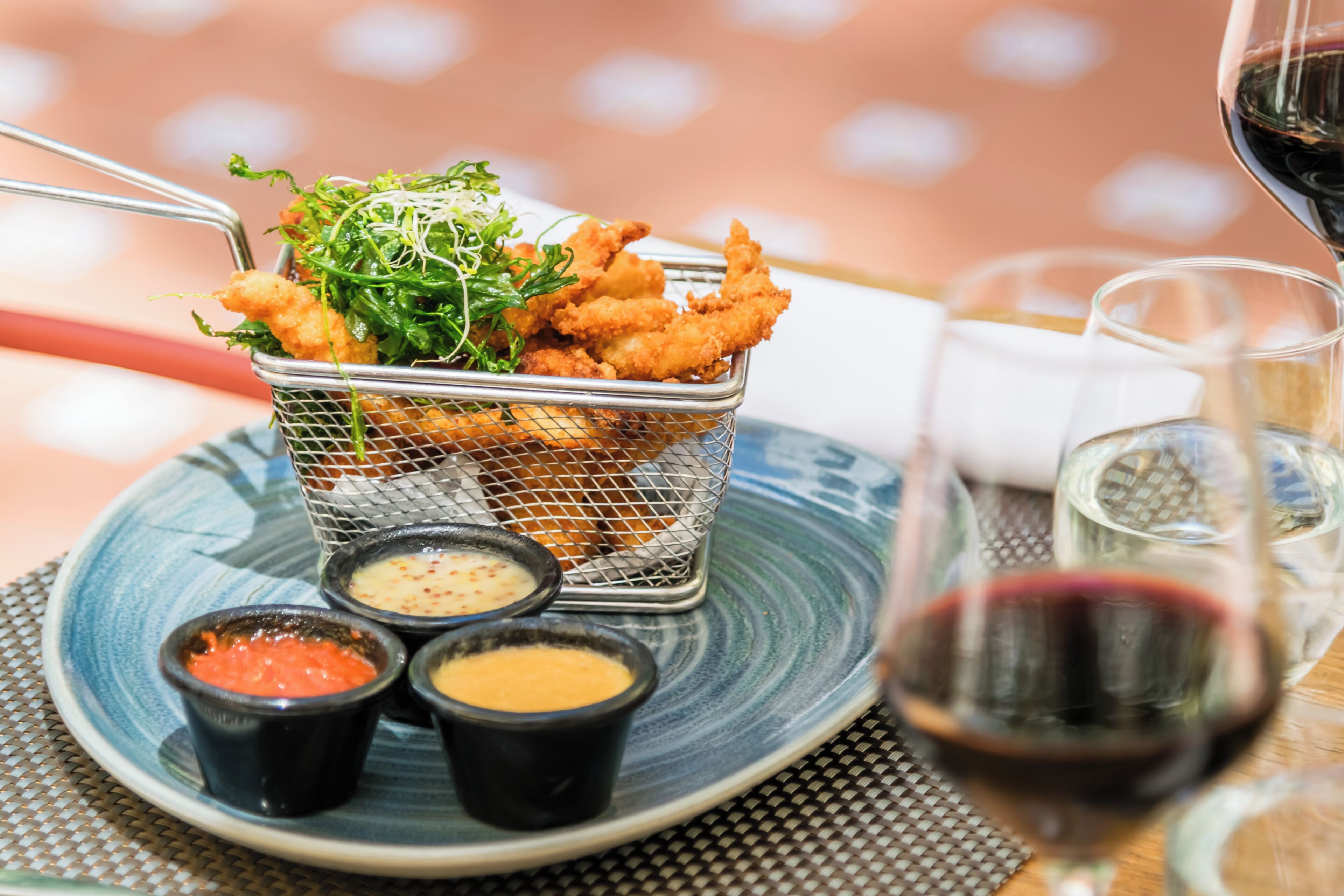 A dining table with a basket of fried food, small bowls of condiments, and a glass of red wine.