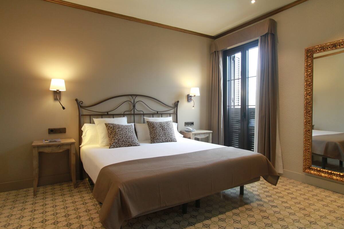 Una habitación de hotel con una cama grande, dos mesitas de noche, un tocador, una ventana con cortinas y un espejo con marco decorativo.