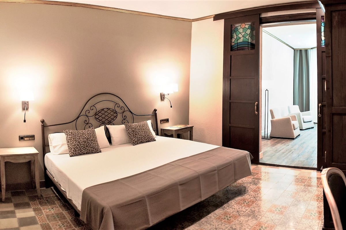 Una habitación de hotel con cama, mesitas de noche, un escritorio con silla y una puerta que da a un balcón.