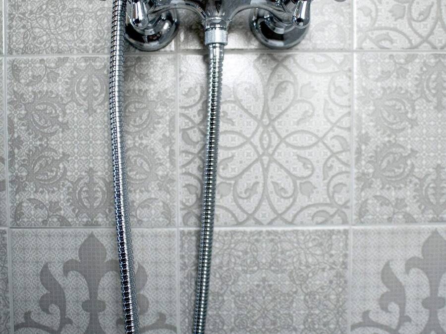 A chrome showerhead with a flexible hose and a white handle against a patterned tile wall.