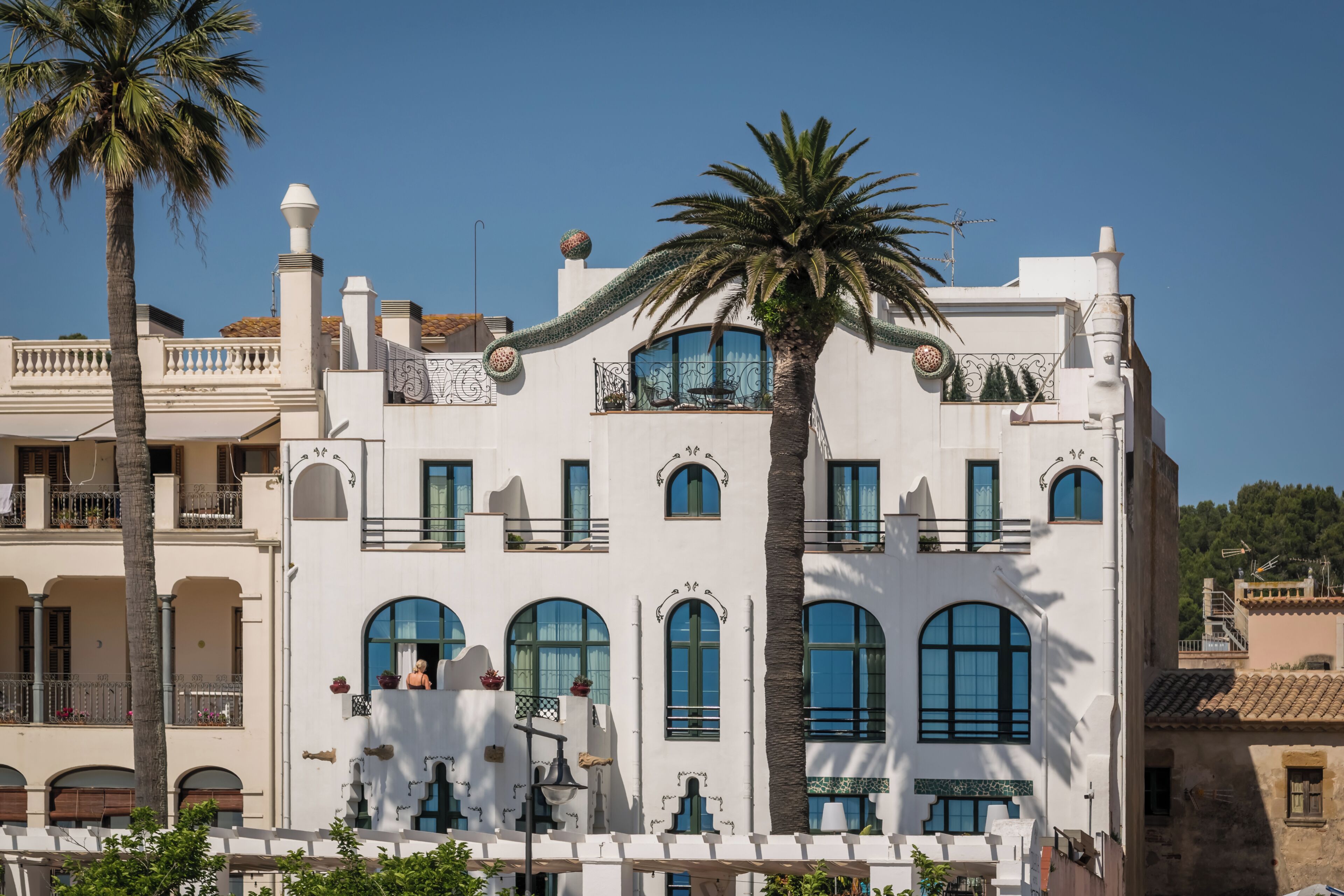 A white building with arched windows and balconies, palm trees, and a clear blue sky.