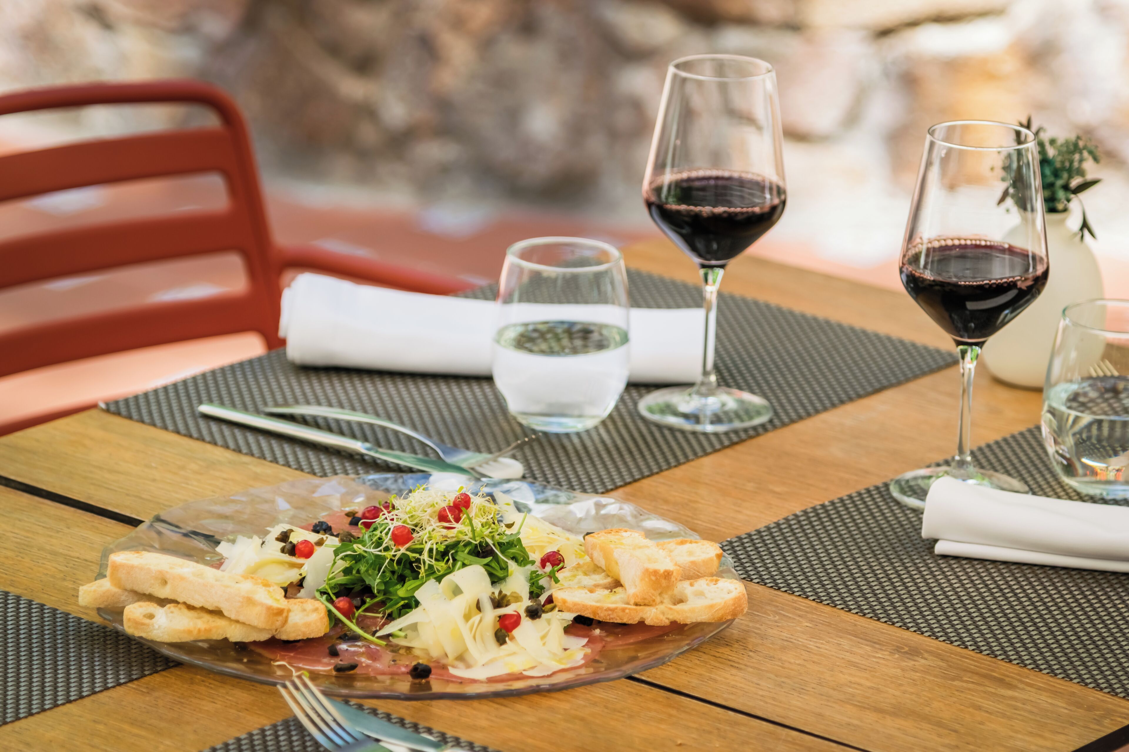 Una mesa de comedor con un plato de comida sobre un vidrio transparente, dos copas de vino y una silla roja.