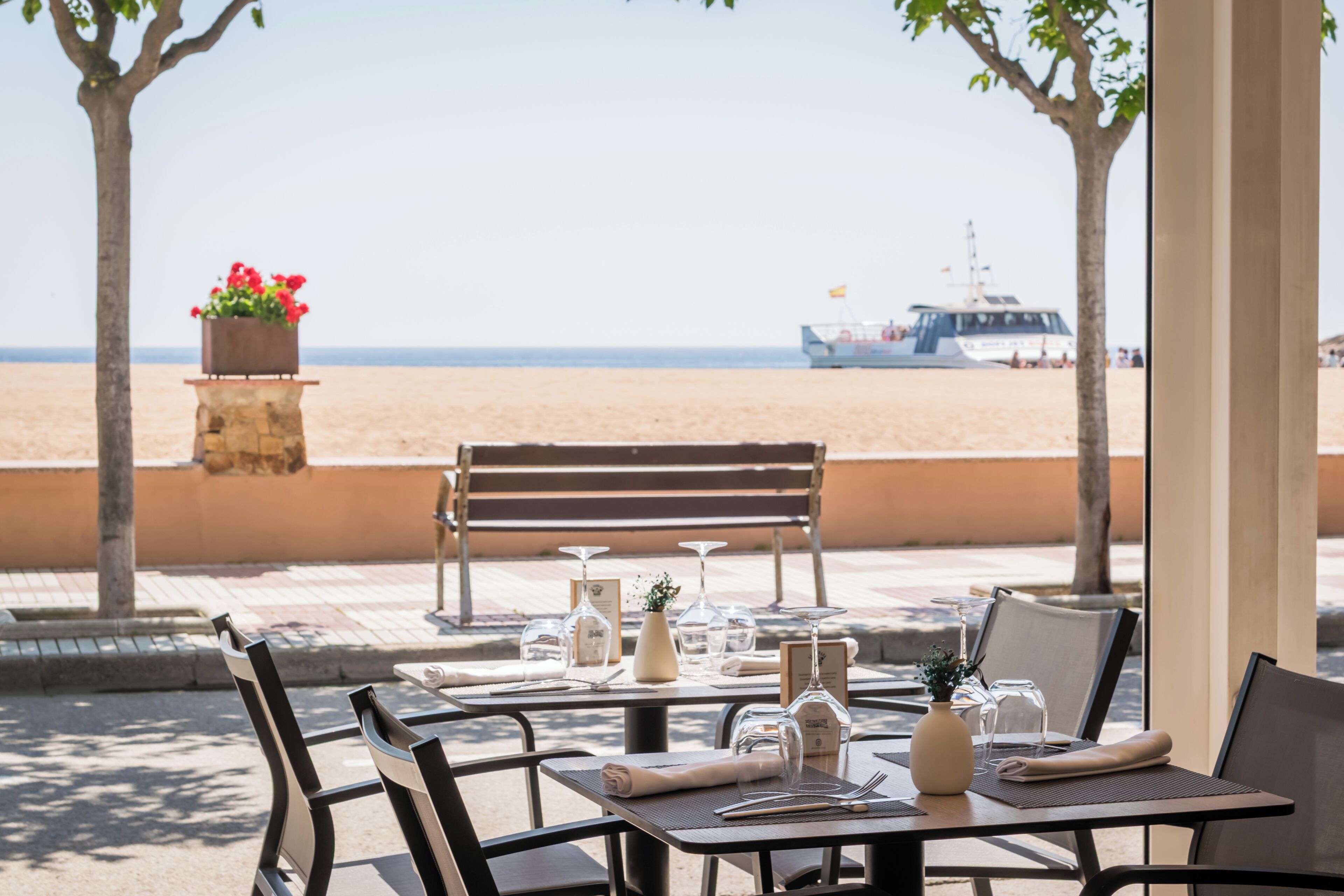 Un espacio de comedor al aire libre con mesas dispuestas para dos, una jardinera de piedra con flores y un banco.
