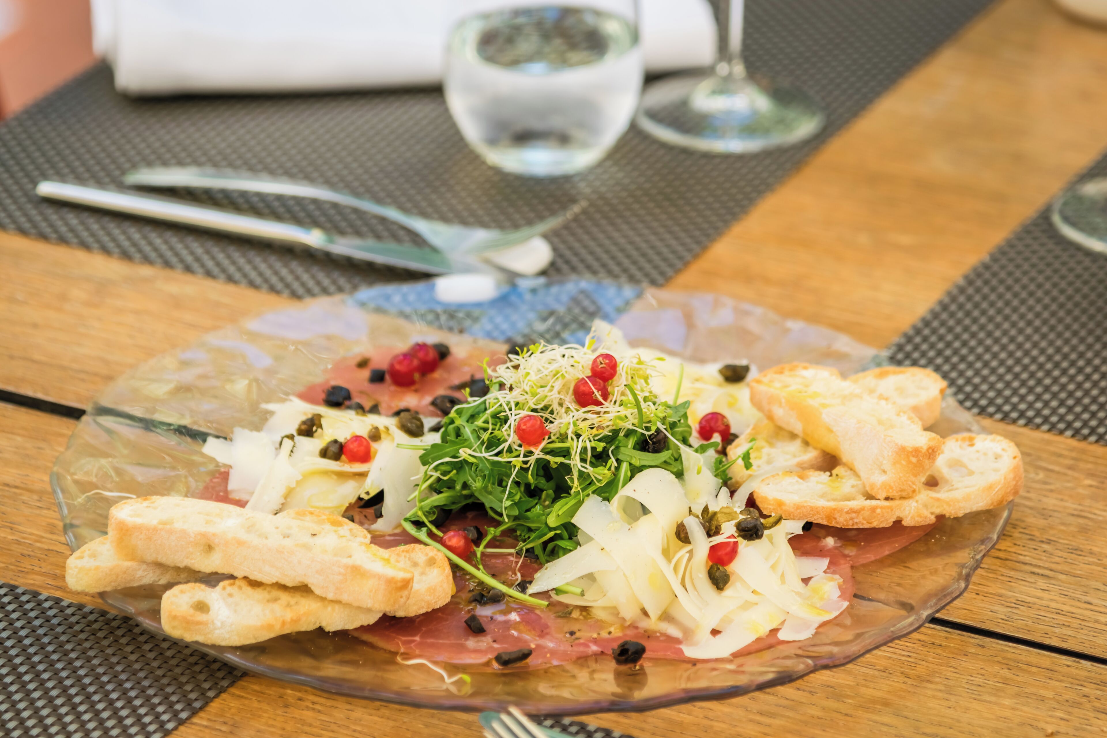 A clear glass plate with a salad, bread, and a water glass on a wooden table.