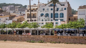 Un pueblo costero con edificios de piedra, palmeras y mesas al aire libre en cafés.