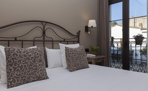 Una cama bien hecha con almohadones estampados, una lámpara de noche y vistas a un balcón con baranda y plantas en macetas.