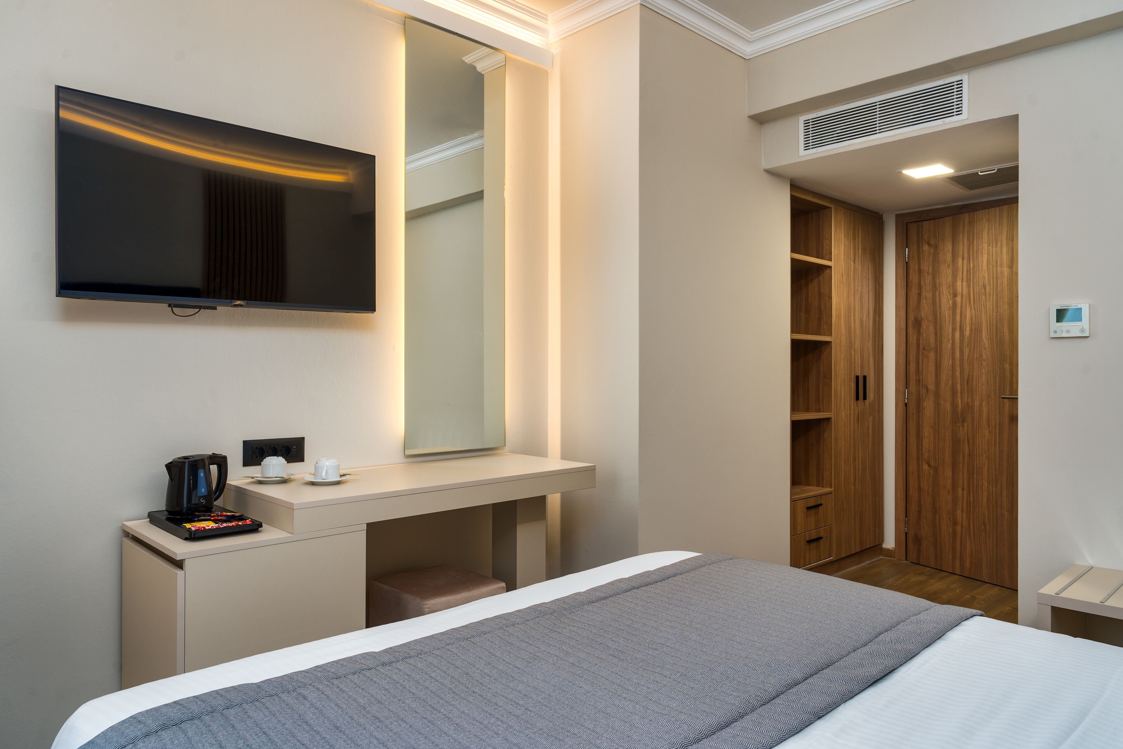 A modern hotel room with a bed, a bedside table with a coffee maker, a flat-screen TV, a mirror, and a wardrobe.