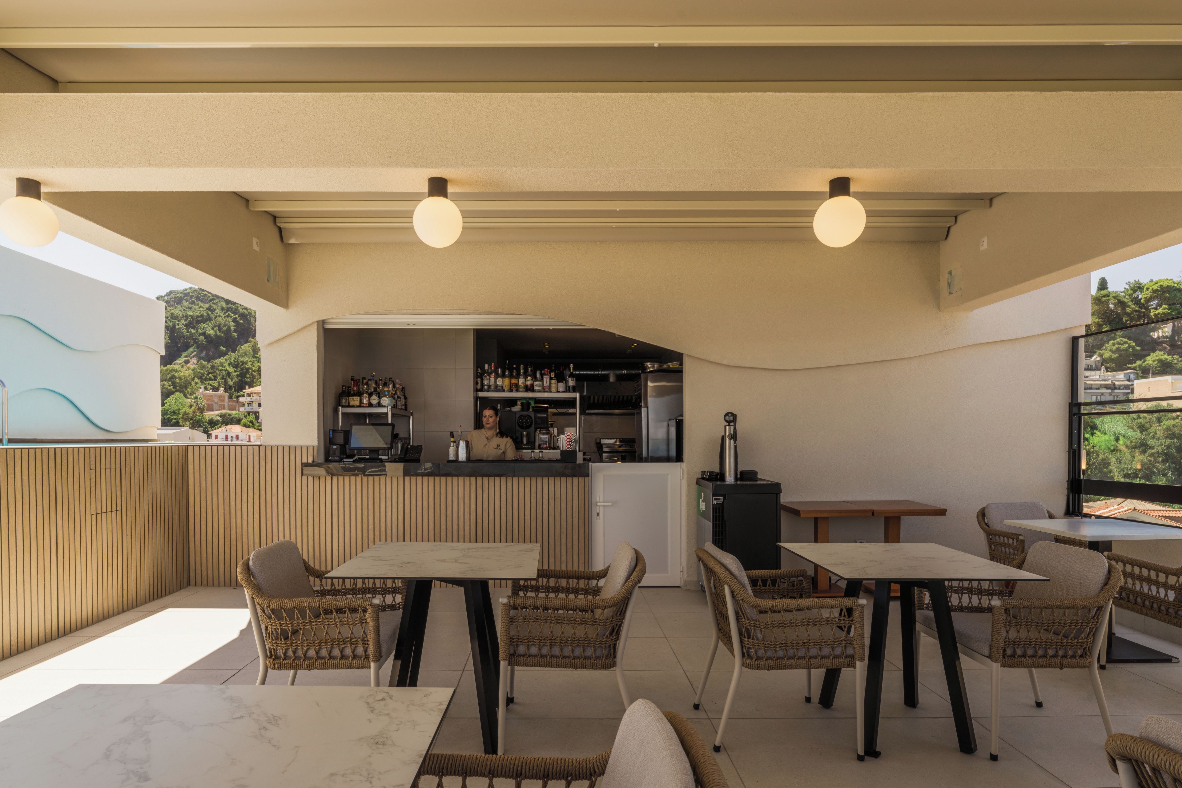 An outdoor seating area with tables and wicker chairs, a bar with a counter and shelves of bottles, and a view of greenery and buildings in the background.