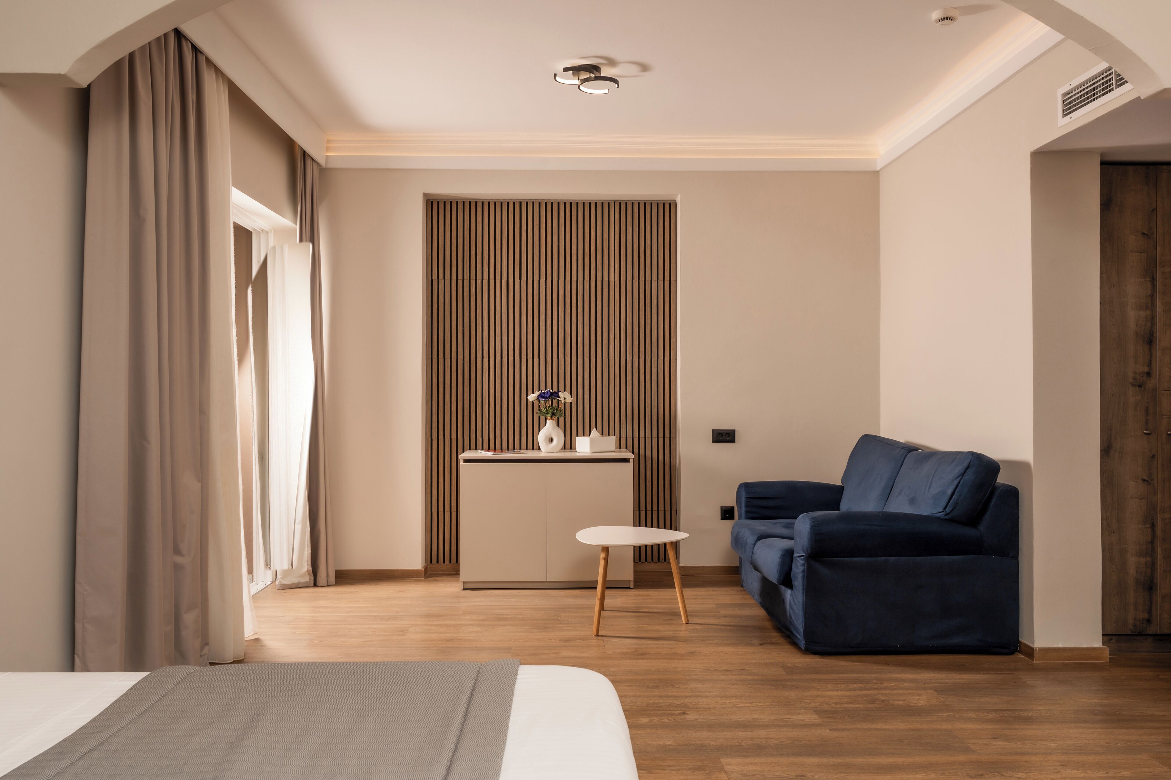 Una habitación de hotel moderna con una cama, un sofá azul, una mesita y una zona de kitchenette.