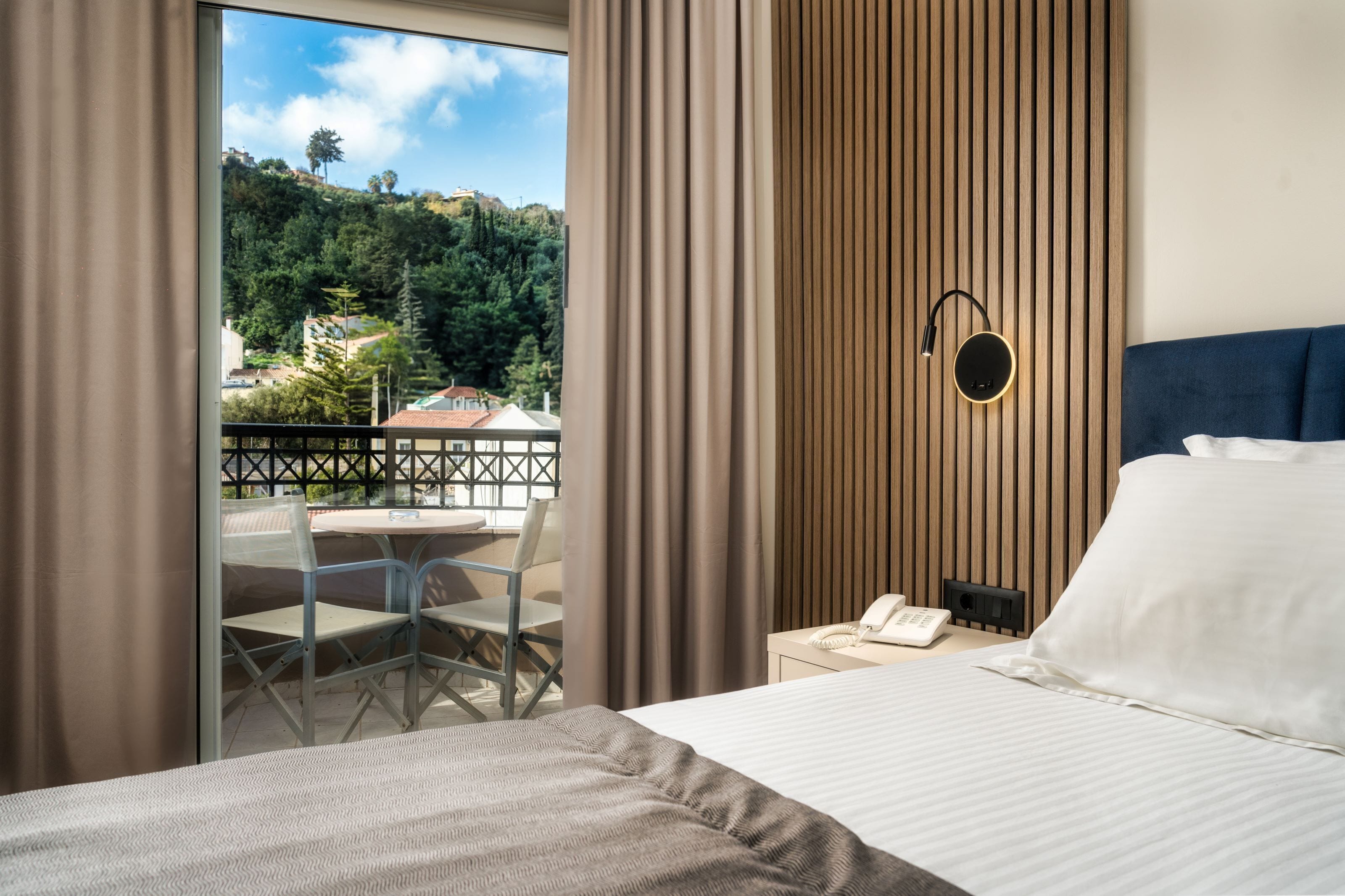 A hotel room with a bed, a desk, a chair, a phone, and a view of a balcony with a table and chairs.