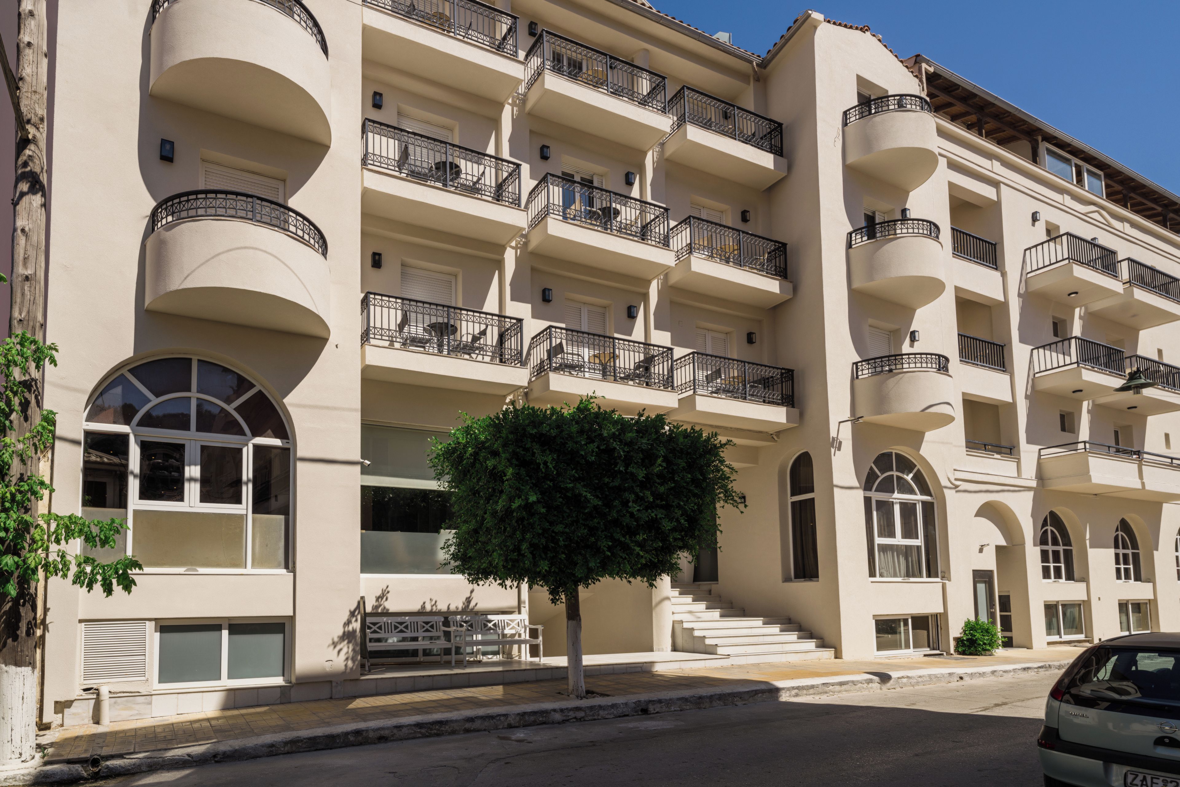 Un hotel de varios pisos con balcones, un árbol en primer plano y un coche estacionado en la calle.