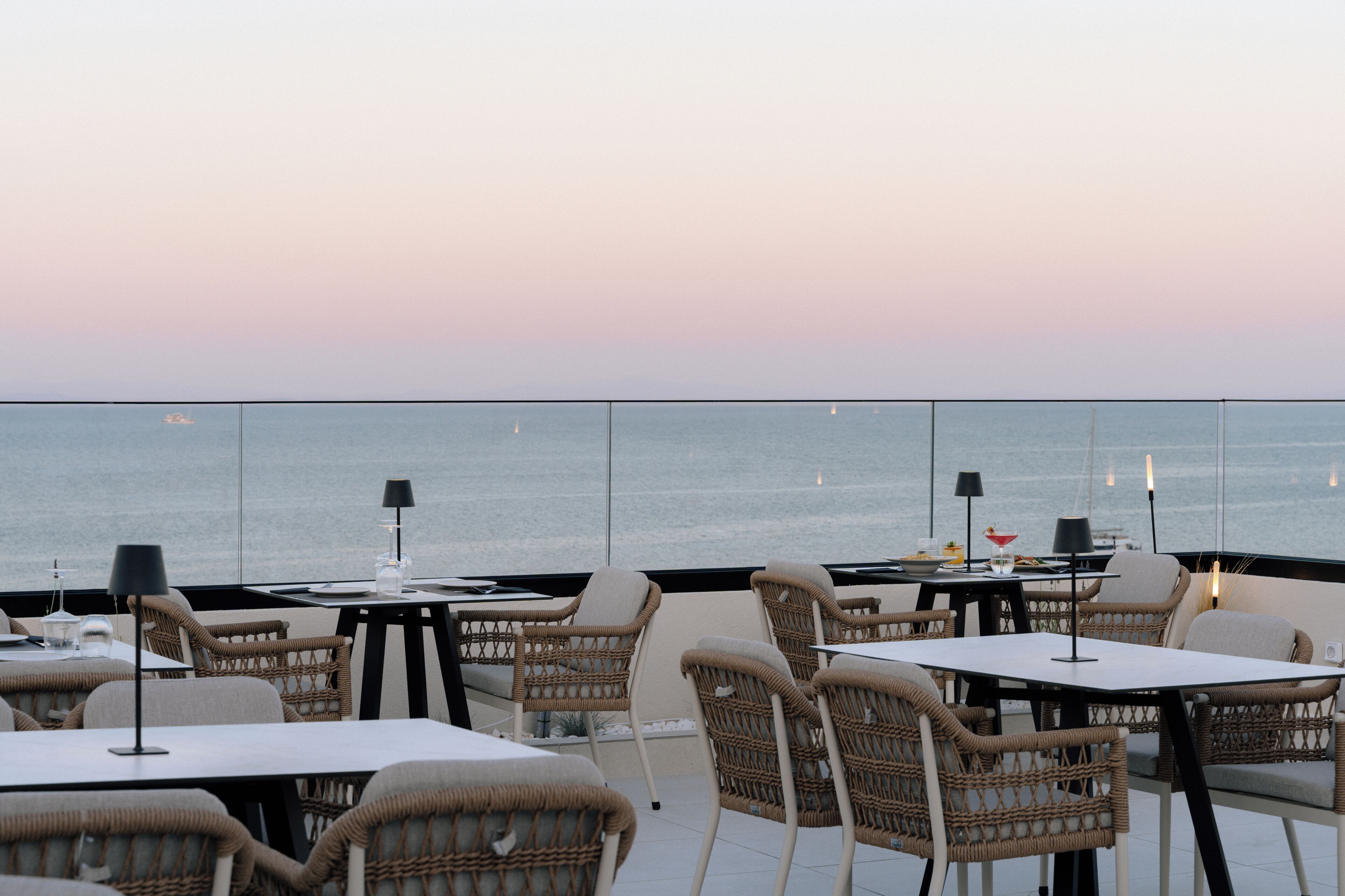 An outdoor dining area with wicker chairs and tables, overlooking the sea.