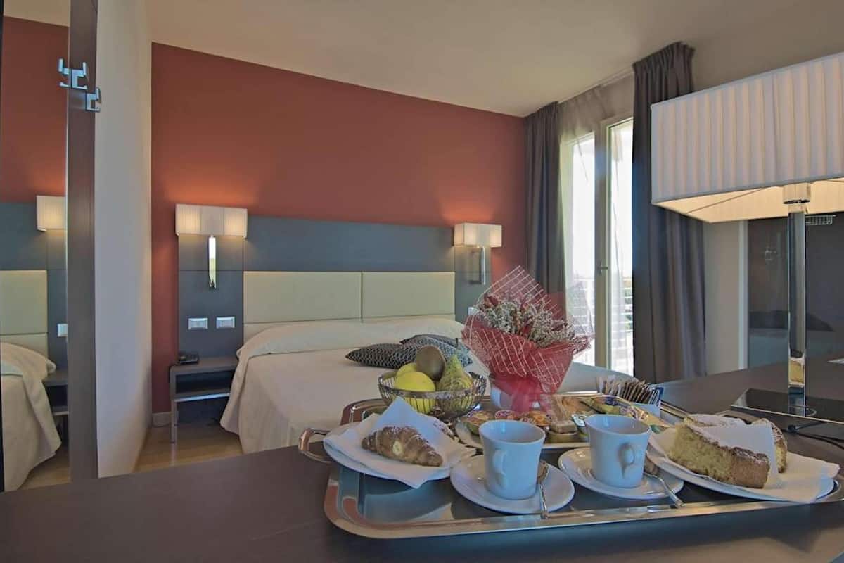 Habitación de hotel con cama, mesitas de noche, una bandeja con alimentos para el desayuno y vistas al exterior.