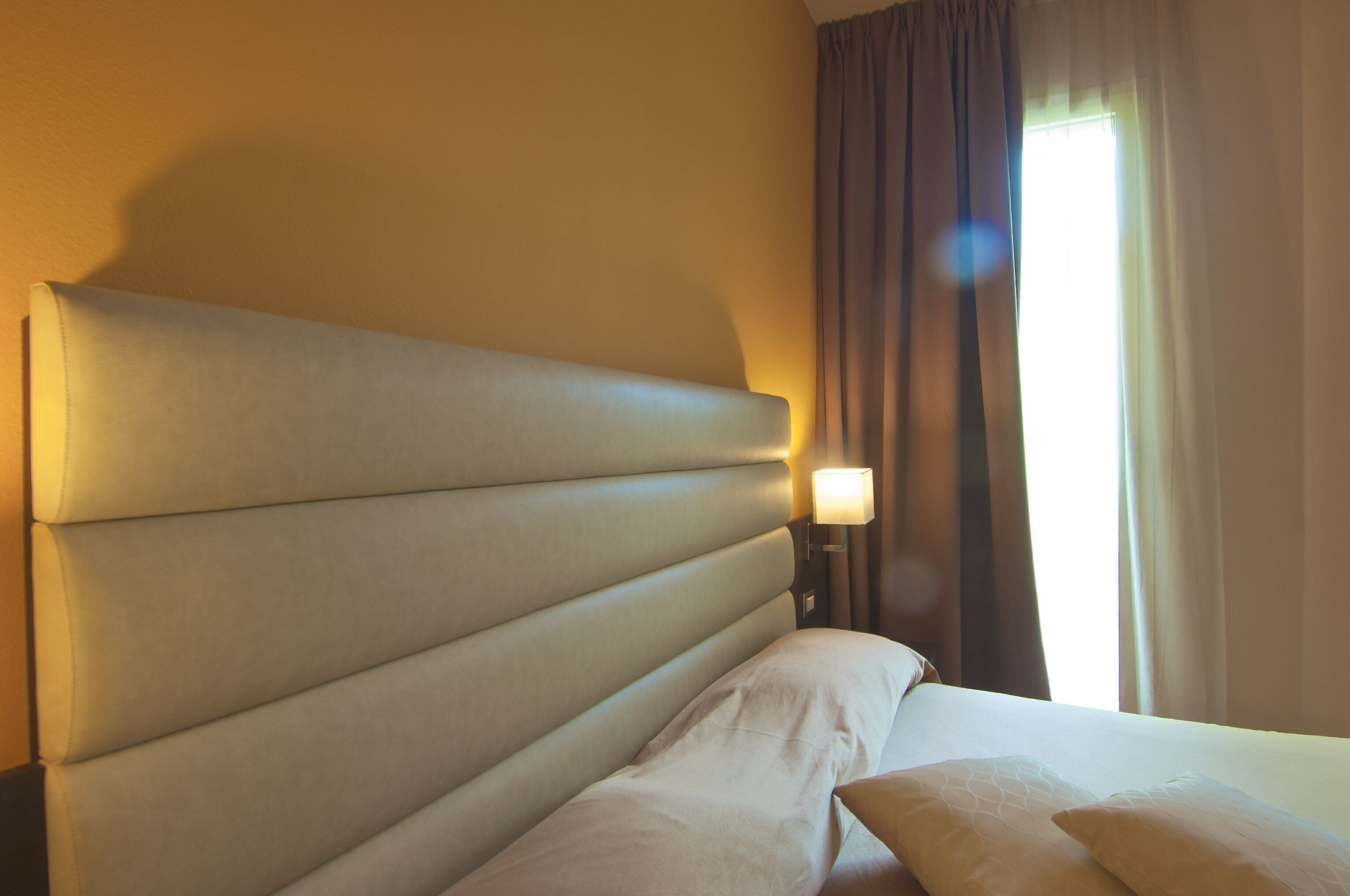 Una habitación de hotel con una cama grande, una mesita de noche, una lámpara y una ventana con cortinas.