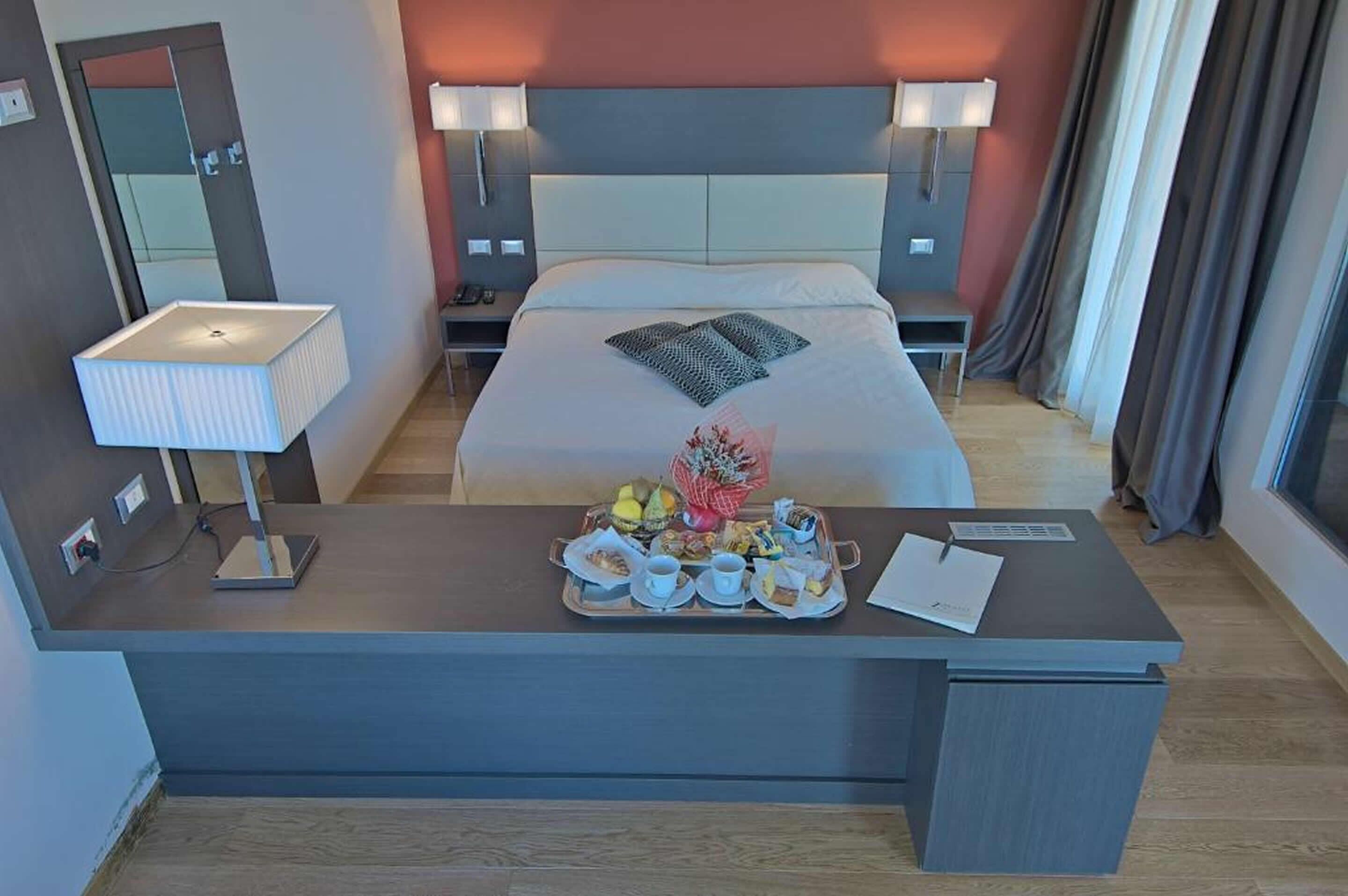A hotel room with a bed, bedside lamps, a desk with a tray of food, a mirror, and a window with curtains.