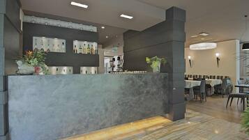 A modern reception area with a marble counter, shelves with bottles, and a dining area in the background.