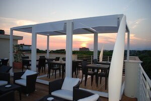 Un patio al aire libre con mesas y sillas, una pérgola blanca y vistas al atardecer sobre un paisaje urbano.