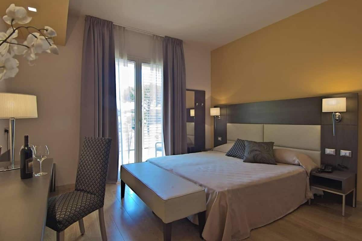 Habitación de hotel con cama, mesitas de noche, una silla, un banco, una lámpara y una ventana con cortinas.