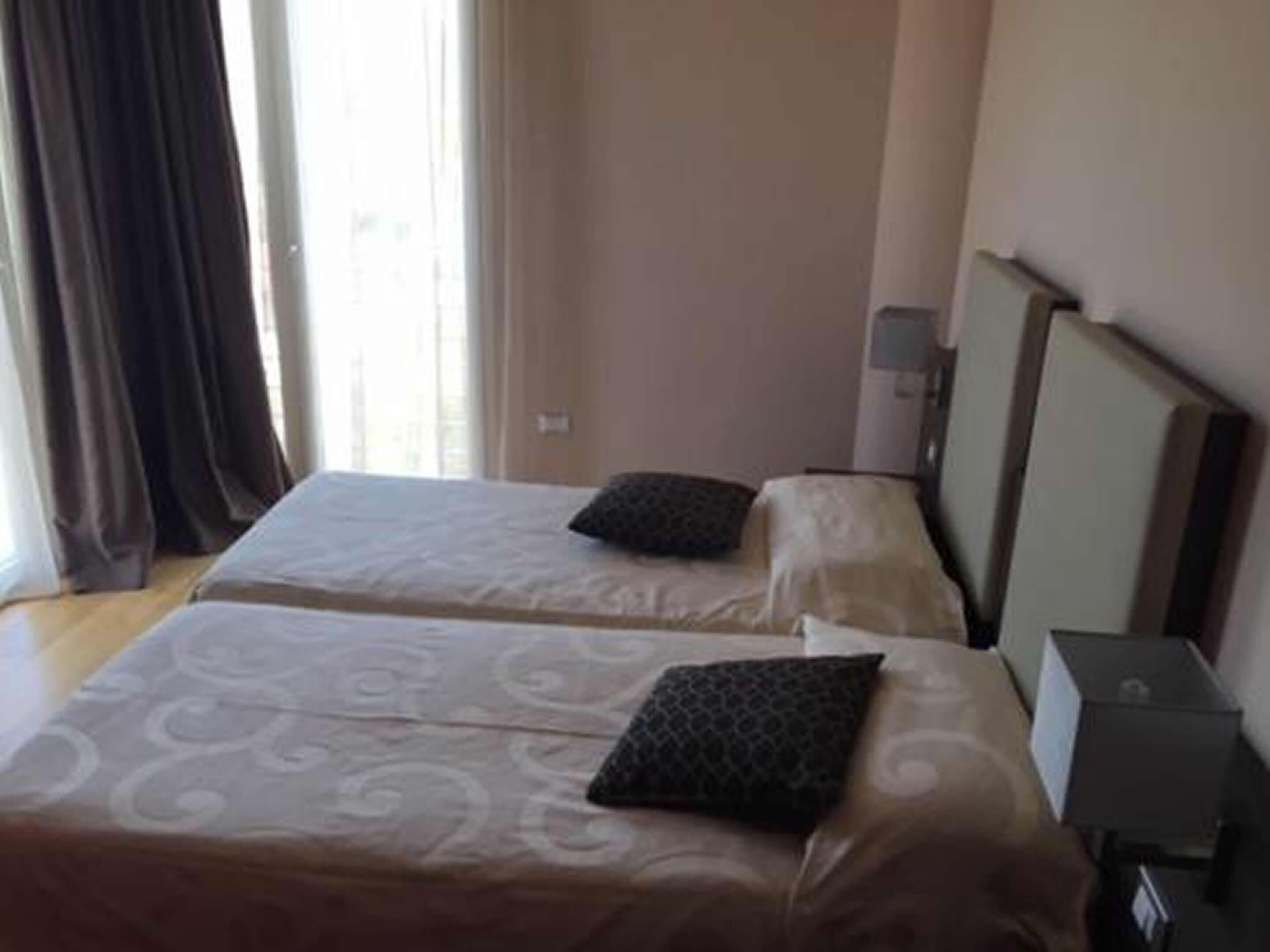 A hotel room with two beds, a nightstand, a lamp, and a window with curtains.