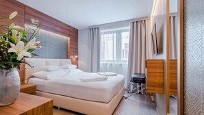 Una habitación de hotel moderna con una cama grande, una mesita de noche, un escritorio pequeño, una silla, una planta en maceta y vistas a la ciudad desde la ventana.
