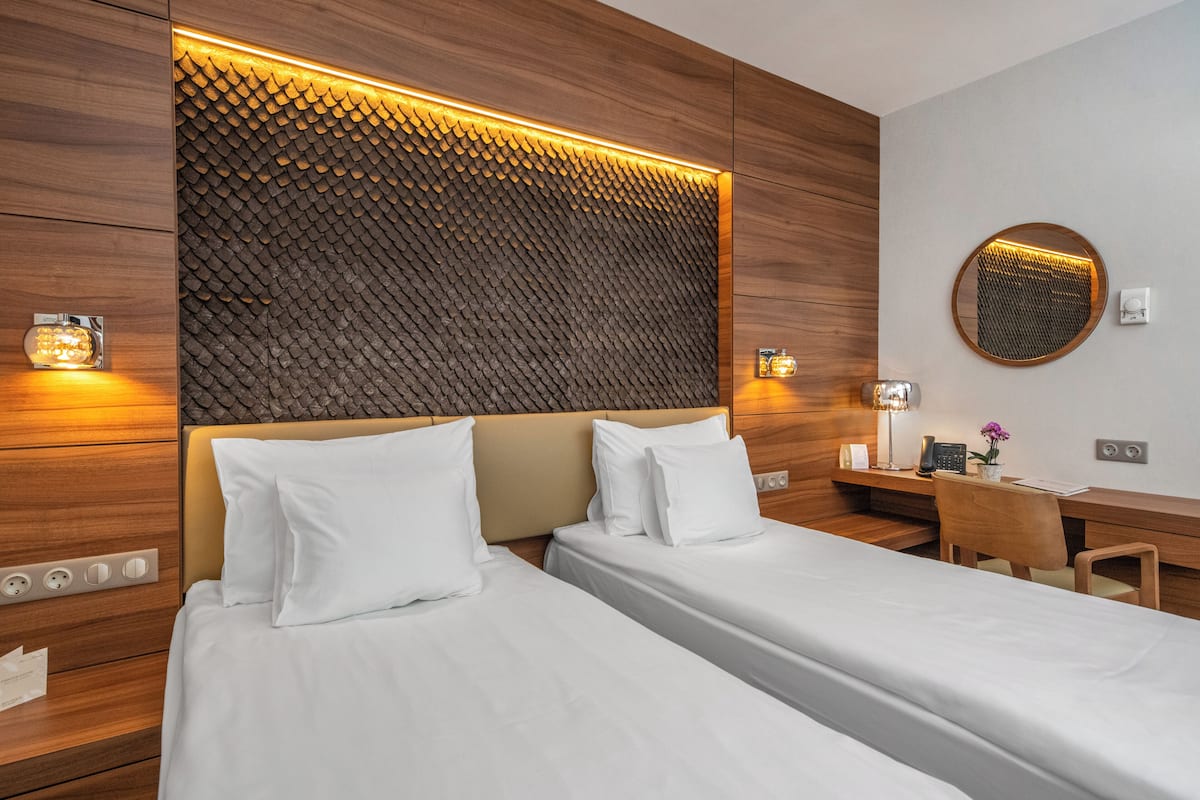 A hotel room with two beds, a wooden headboard, a desk, and a round mirror.
