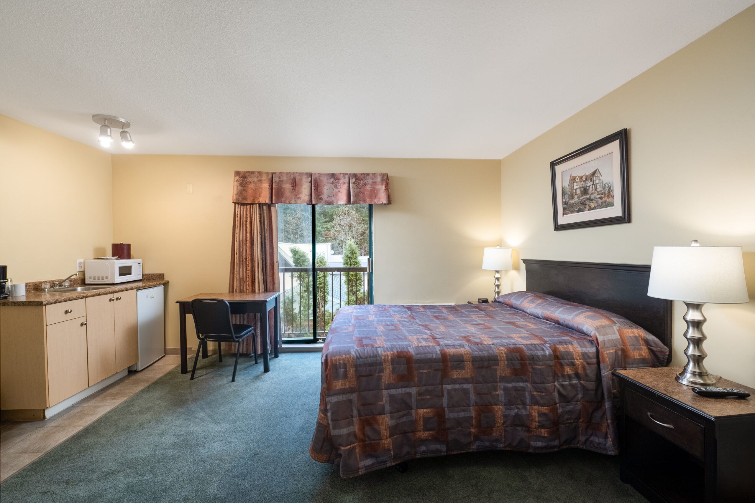 A hotel room with a bed, a desk with a chair, a microwave, a lamp, and a window with curtains.