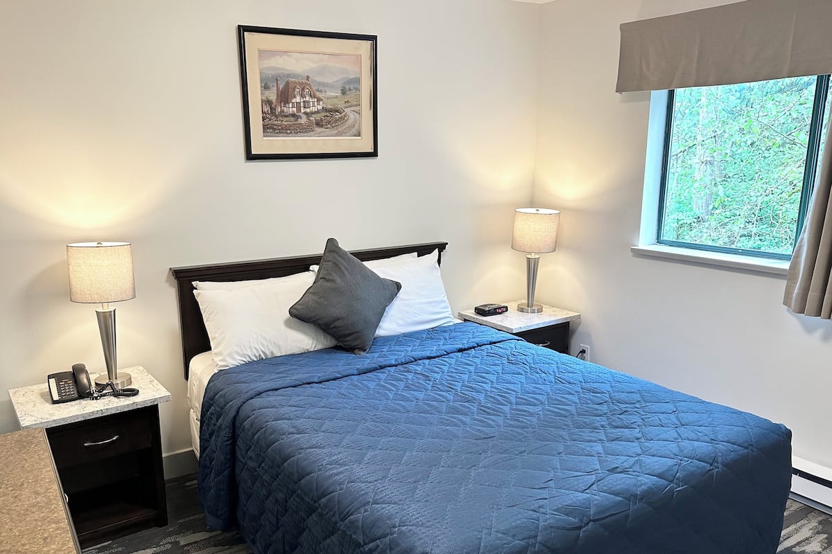 A neatly made bed with a blue quilt, two bedside tables with lamps, a framed picture on the wall, and a window with a view of trees.