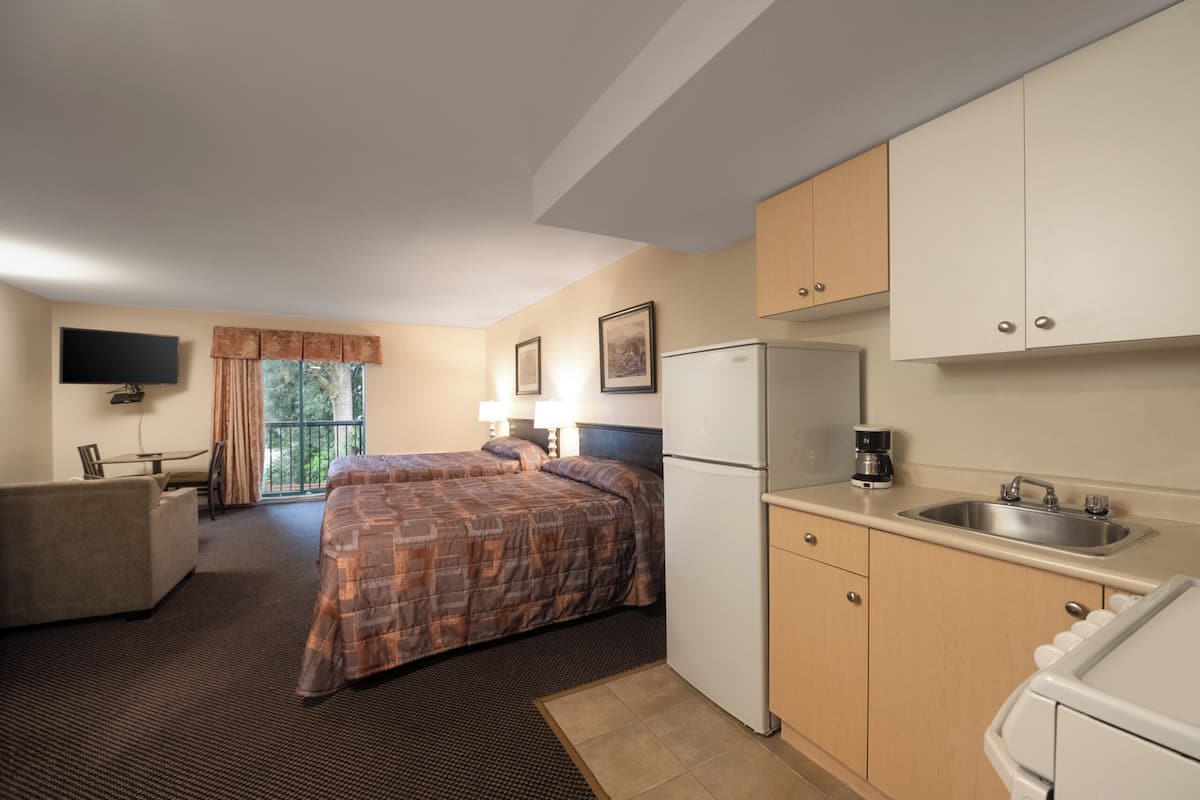 A compact hotel room with a bed, a small kitchenette, a flat-screen TV, and a window with curtains.