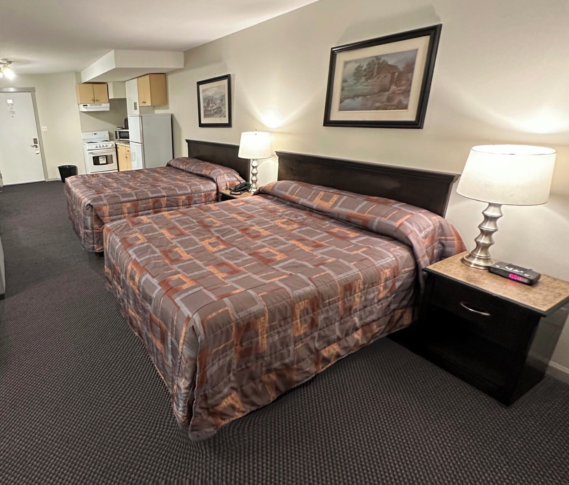 A hotel room with two beds, a kitchenette, and a painting on the wall.