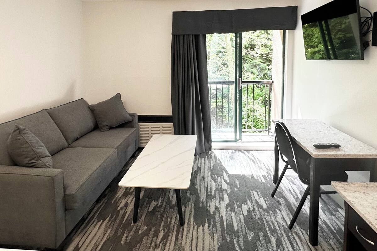 A modern living room with a grey sofa, a white coffee table, and a small desk with a chair. There is a large window with curtains, and a TV mounted on the wall.