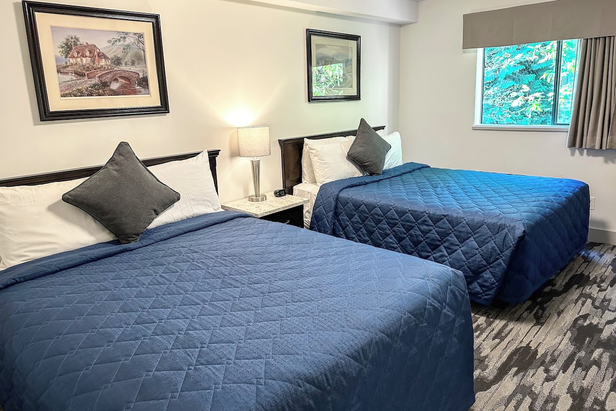 A hotel room with two beds, each with a blue quilted bedspread and a pillow. There are framed pictures on the wall, a nightstand with a lamp, and a window with curtains.