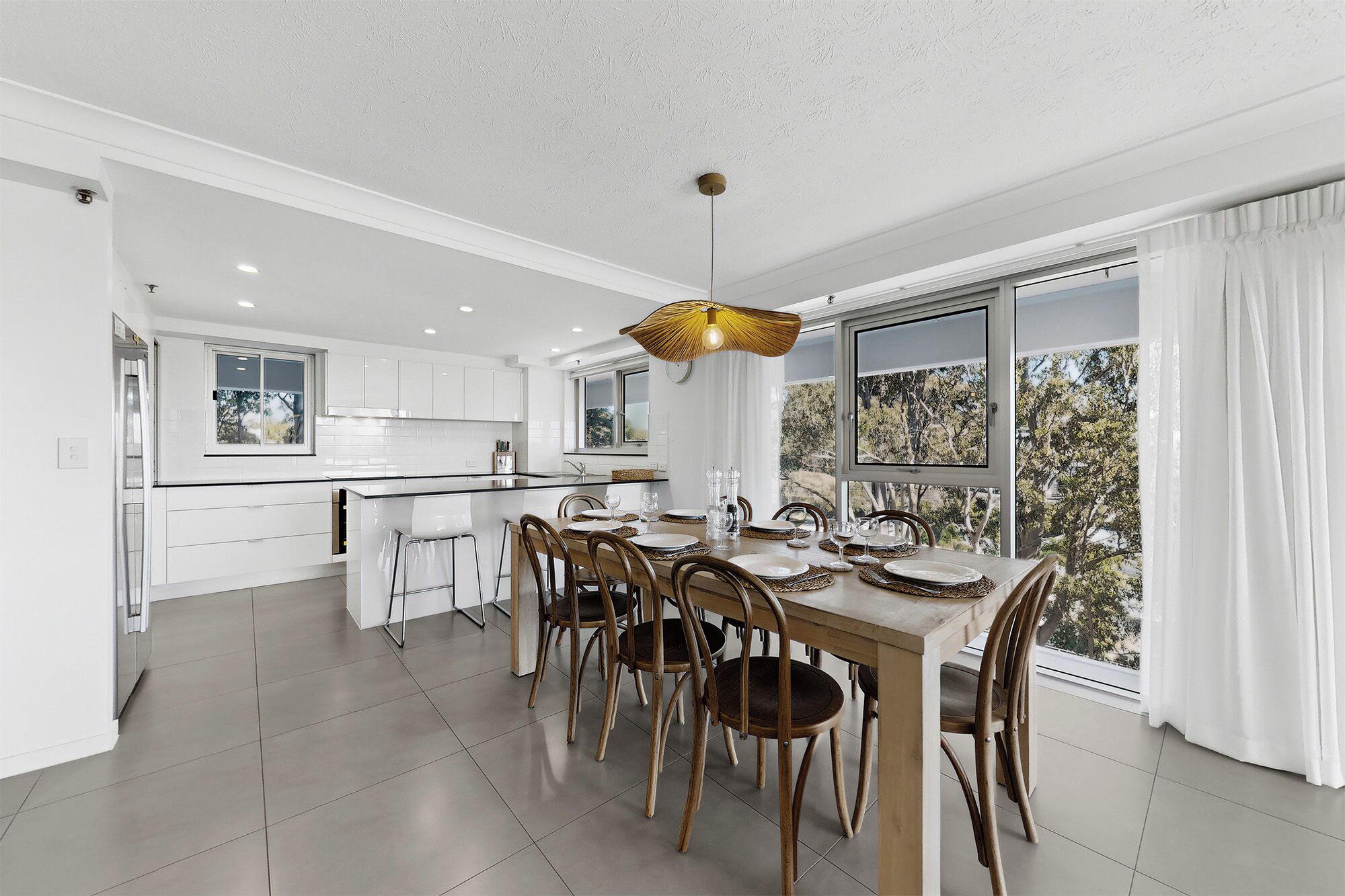Une cuisine moderne avec des meubles blancs, un coin repas comprenant une table et des chaises en bois, et de grandes fenêtres offrant une vue sur les arbres.