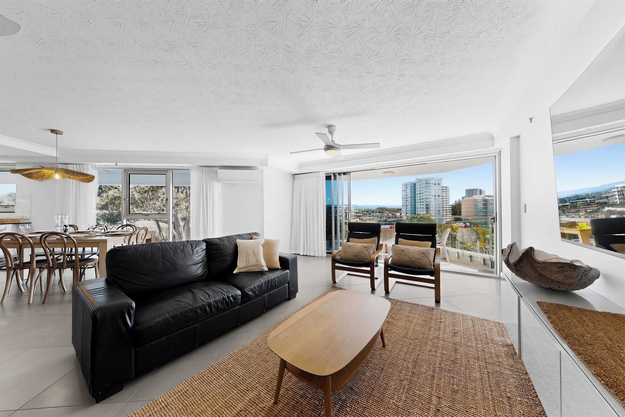 A modern living room with a black leather sofa, a wooden coffee table, and a view of the city through large windows.