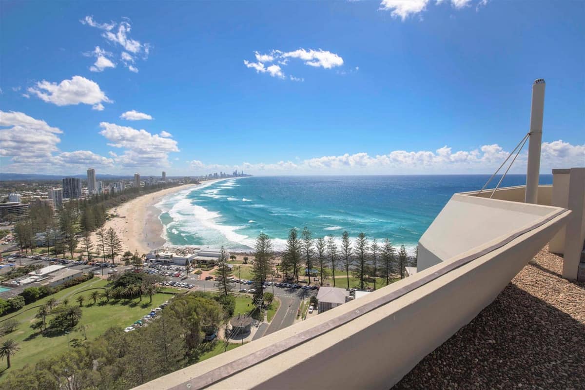A coastal view from a balcony overlooking a beach and ocean, with buildings and a parking lot in the foreground.