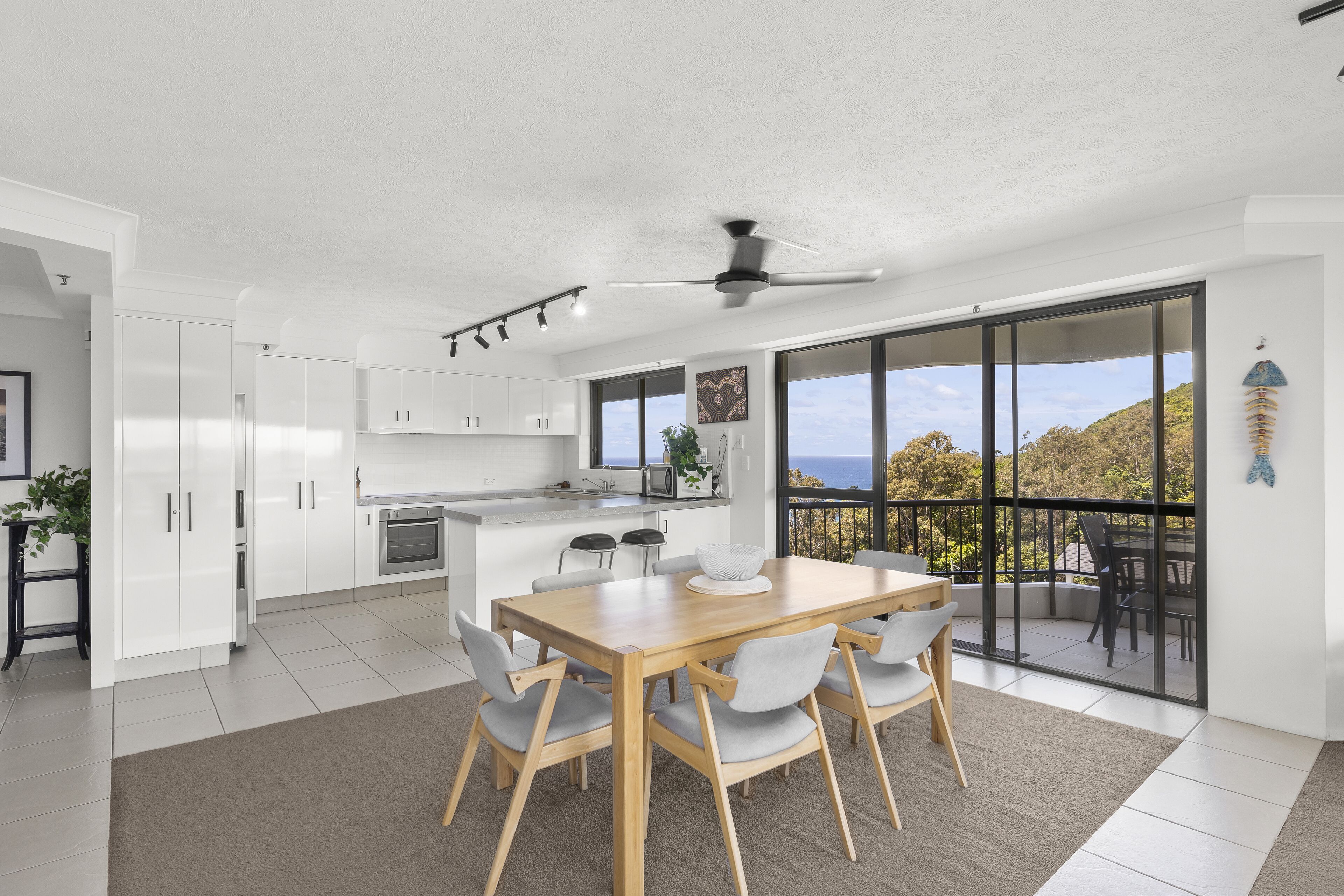 A modern kitchen with white cabinets, a wooden dining table, and a large sliding glass door leading to a balcony with a view of the hills.
