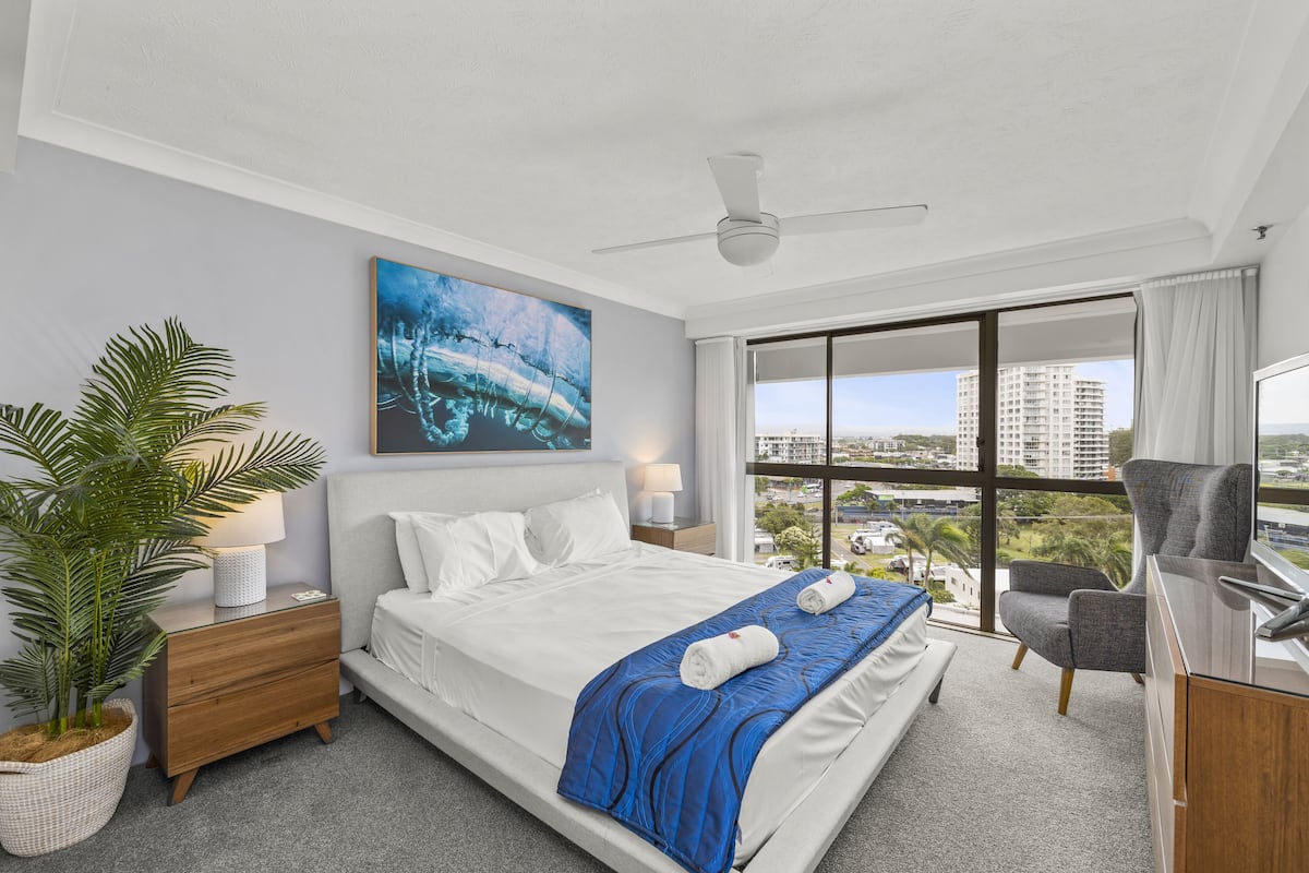 A modern hotel room with a large bed, a desk, a chair, a large window with a city view, and a ceiling fan.