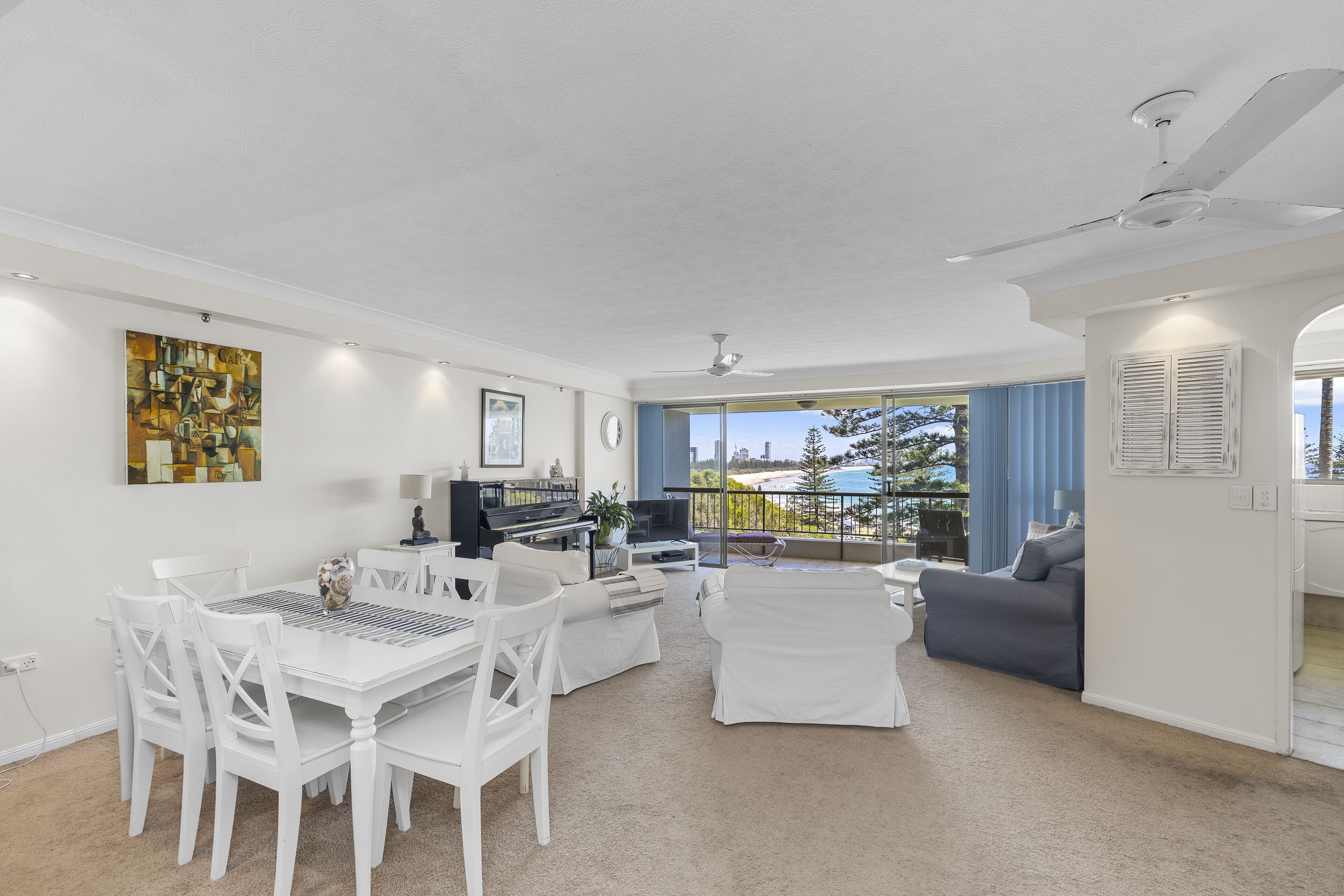 A spacious living area with a dining table, white chairs, a gray sofa, and a large window offering a view of the outdoors.