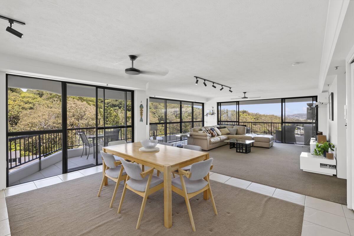 A spacious living area with a wooden dining table and chairs, a sofa, and a balcony with a view of greenery.
