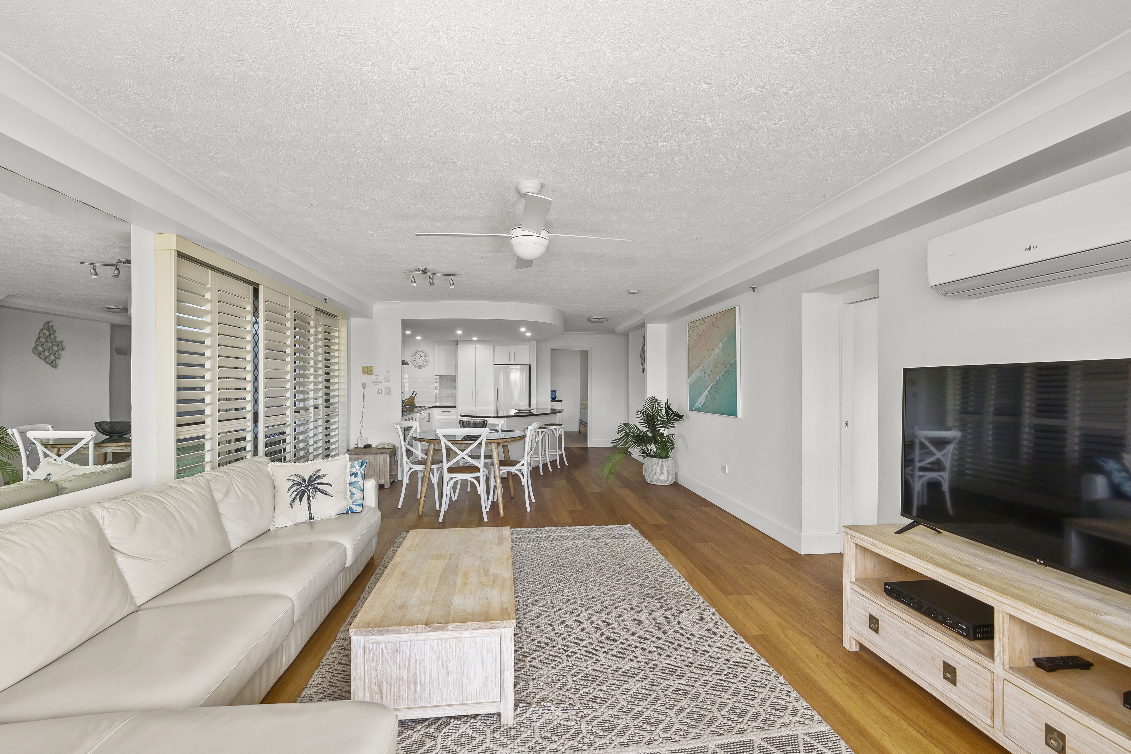 A modern living room with a white sofa, a wooden coffee table, and a TV on a wooden stand. There is a dining area in the background with white chairs and a kitchen visible.