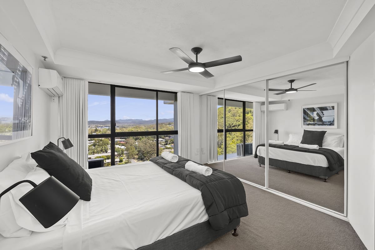 A modern bedroom with a large bed, a ceiling fan, and a view of the city through large windows.